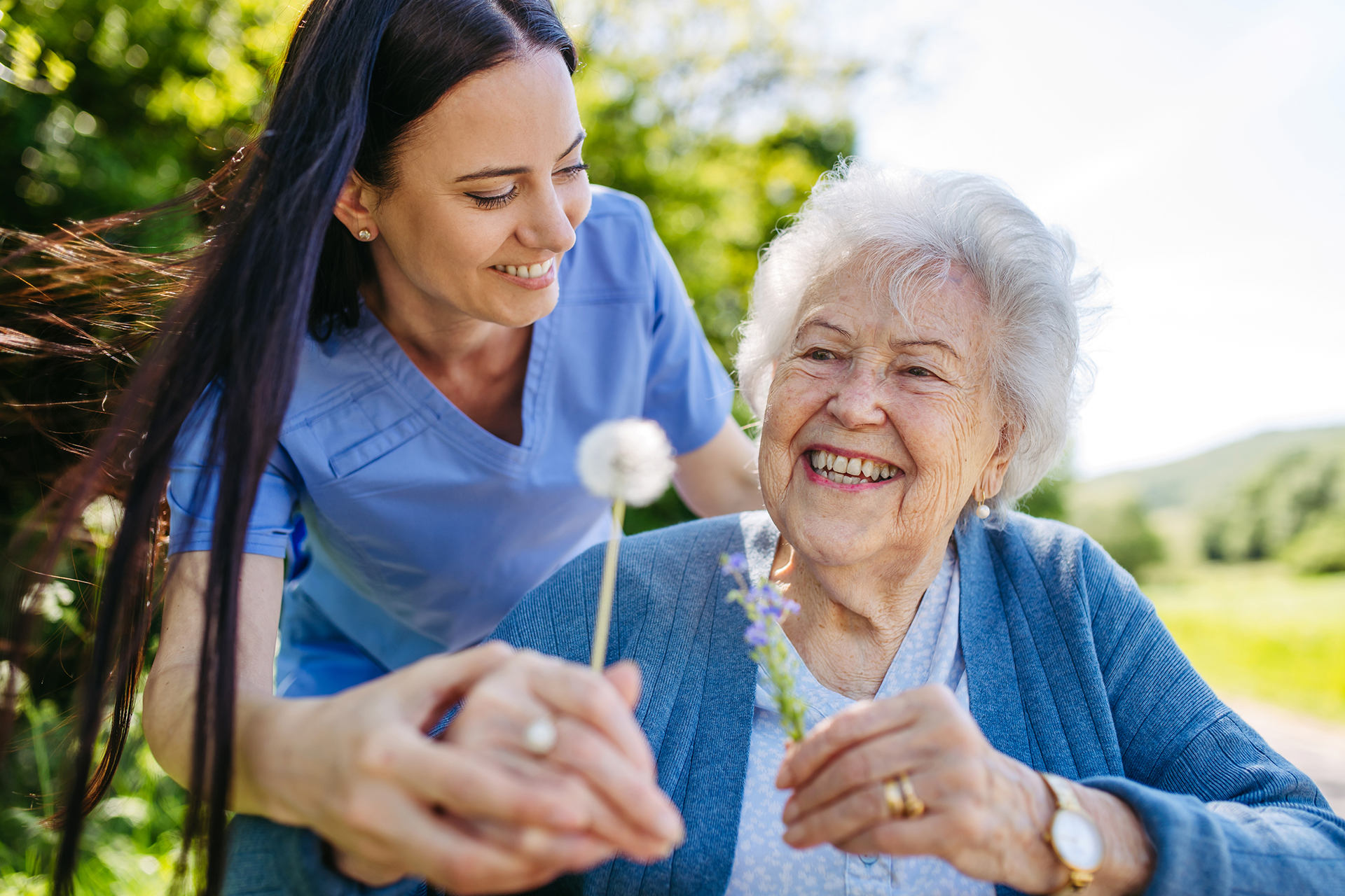 Caregiver assisting an older adult with a dandelion during a joyful outdoor moment.