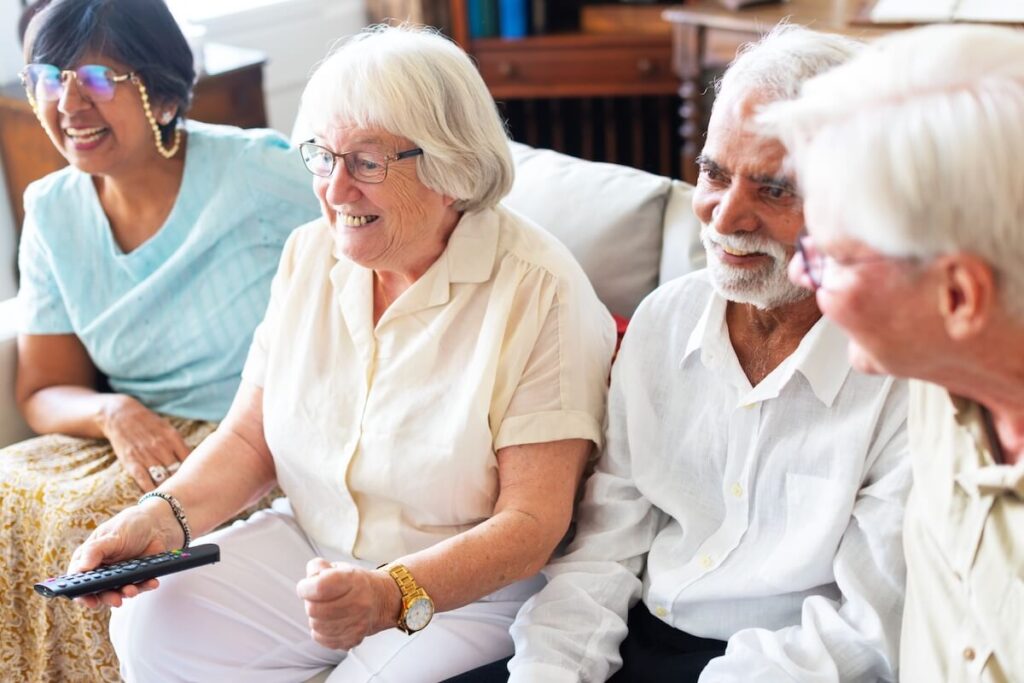 Group of diverse senior friends sitting on a couch, smiling and watching TV together.