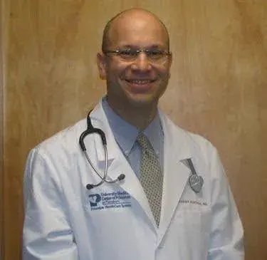 A man wearing a white coat and stethoscope standing against a wooden background, smiling.