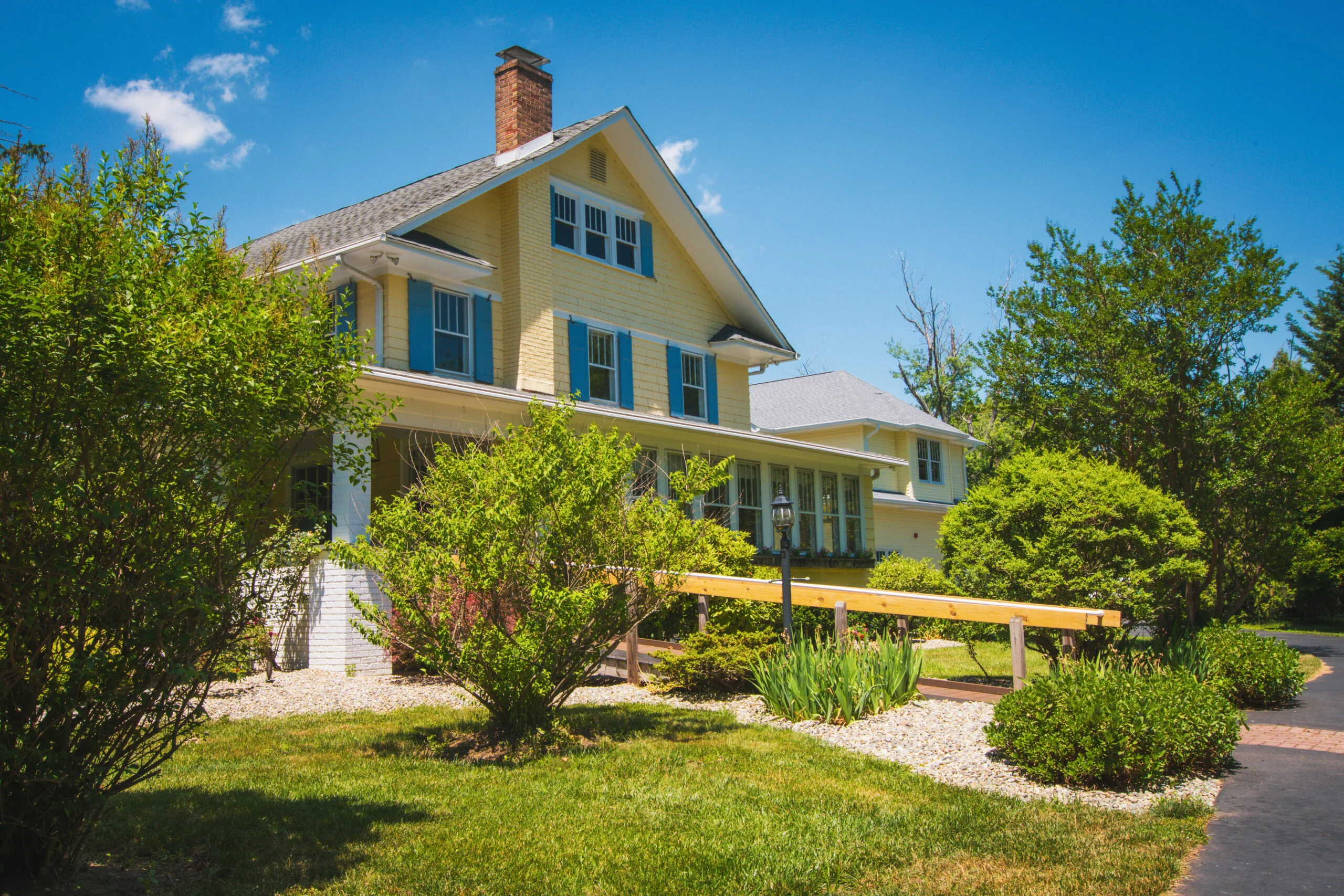 Yellow two-story senior living community building with blue shutters surrounded by green bushes, trees, and a grassy lawn with a paved pathway and wooden handrail leading to the entrance under sunny daylight.