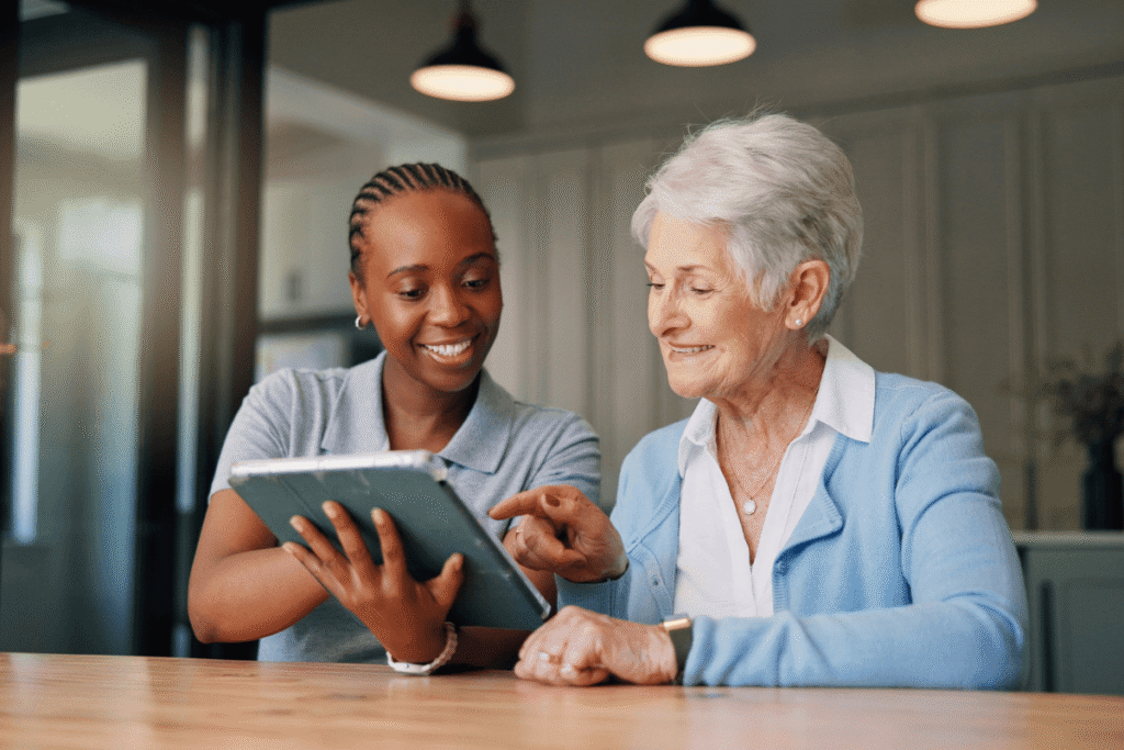 A caregiver and an elderly woman share a joyful moment over a tablet, celebrating positive news, medical results, or a successful telehealth appointment at home.